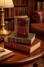 Load image into Gallery viewer, Stack of old books on a wooden table with a lamp and chair in the background