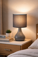Load image into Gallery viewer, Gray table lamp on a nightstand next to a bed with a wooden headboard.