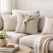 Load image into Gallery viewer, Beige sofa with decorative pillows featuring a goat design, a blanket, and a small table with a vase and books.