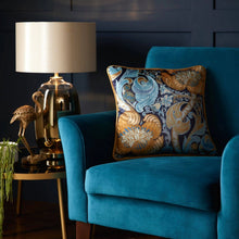 Load image into Gallery viewer, Blue armchair with a decorative pillow next to a lamp on a side table against a dark wall.