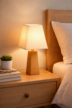 Load image into Gallery viewer, Wooden lamp on a nightstand next to a bed with pillows