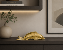 Load image into Gallery viewer, A gold decorative fish sculpture displayed on a dark wooden console table in a modern interior setting with a beige wall, framed art, a shelf with books, and a vase with greenery.
