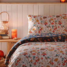Load image into Gallery viewer, Bed with autumn-themed bedding featuring animals and nature patterns, next to a wooden nightstand with a candle and books.