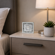 Load image into Gallery viewer, A square white alarm clock with a black Roman numeral face and a decorative dark border sits on a light wood nightstand beside a lit table lamp and a small potted succulent.