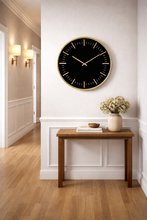 Load image into Gallery viewer, Black wall clock on a white wall above a wooden table with a vase of flowers.