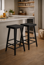 Load image into Gallery viewer, Two black bar stools in a kitchen setting with a countertop and shelves in the background.