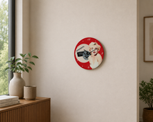 Load image into Gallery viewer, A circular wall clock featuring a red background and a Marilyn Monroe design, mounted on a beige wall in a modern room setting with a wooden cabinet and plants.