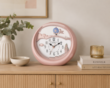 Load image into Gallery viewer, A round pink wall clock with a whimsical illustrated face sits on a light wood console table in a styled room setting, next to a vase with greenery, a small bowl, and books.