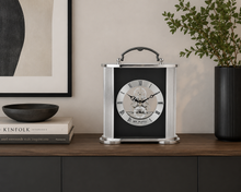 Load image into Gallery viewer, A silver and black table clock with a handle and skeleton dial, displayed on a wooden shelf next to a bowl, books, and a potted plant.