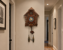 Load image into Gallery viewer, A carved wooden cuckoo clock with a detailed house-shaped frame hangs on a beige hallway wall. The clock features a circular clock face with Roman numerals, a small window, and dangling pinecone-shaped weights.