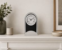 Load image into Gallery viewer, A black and silver mantel clock with a white face and Roman numerals, styled on a white mantelpiece with a vase, candle, and bowl against a neutral wall.
