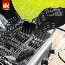 Load image into Gallery viewer, A black heat-resistant grill cleaning glove with gray grip dots and a silver metal wire brush is shown cleaning a charcoal grill outdoors on a wooden deck with green grass in the background. The 'Let's BBQ' logo is visible in the top-left corner.
