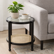 Load image into Gallery viewer, Round black side table with glass top and wicker shelf, featuring a plant and tea cup on a living room floor.
