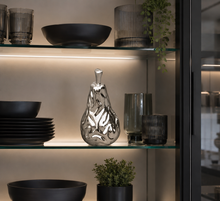 Load image into Gallery viewer, A polished silver pear-shaped decorative vase with a leaf-like cutout pattern, displayed on a lit glass shelf alongside black bowls, dark glass tumblers, and small potted plants.
