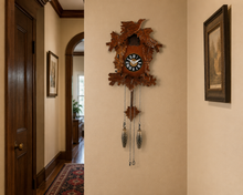 Load image into Gallery viewer, A carved wooden cuckoo clock with leaf and bird details hangs on a beige wall in a hallway with wood trim, framed artwork, and a patterned rug.

