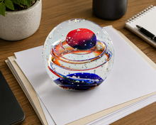 Load image into Gallery viewer, A clear glass decorative orb with swirling red, blue, and orange patterns sits on stacked white papers on a wooden desk, with a plant, notebook, and pen nearby.
