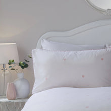 Load image into Gallery viewer, White bed with heart-patterned pillows next to a nightstand with a lamp and decorative items.
