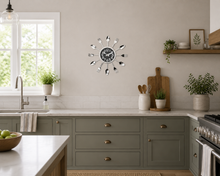 Load image into Gallery viewer, A modern wall clock with a black round face and silver spoon-shaped hands, mounted on a light beige wall in a styled kitchen setting with sage green cabinets, white subway tile, and potted plants.
