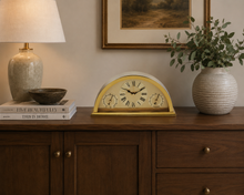 Load image into Gallery viewer, A vintage-style gold and cream mantel clock with Roman numerals sits on a dark wood sideboard in a beautifully styled room, flanked by a lamp and a potted eucalyptus plant.

