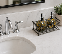 Load image into Gallery viewer, Two round mosaic soap and lotion dispensers with black and gold patterns sit in a black metal tray on a white marble bathroom counter next to a chrome faucet.
