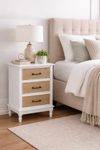 Load image into Gallery viewer, White nightstand with wicker drawers next to a bed with beige headboard and pillows.
