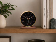 Load image into Gallery viewer, A round analog clock with a black face, gold-tone numerals and hands, and a gold outer rim sits on a wooden shelf. The shelf is styled with a potted green plant on the left and a stack of books on the right, including 'KINFOLK' and 'EAT DRINK NAP'.
