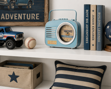 Load image into Gallery viewer, A light blue retro-style radio with a clock face on the front, styled on a white shelf alongside books, a toy truck, a baseball, and a striped pillow in a home decor setting.
