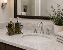 Load image into Gallery viewer, A bathroom vanity countertop with a white oval sink, polished chrome faucet and handles, two patterned soap dispenser bottles in a black wire rack, a large dark wood mirror, a wall sconce, folded white towels, and a potted eucalyptus plant.
