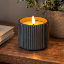 Load image into Gallery viewer, Candle in a textured black holder on a wooden surface with a plant in the background
