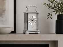 Load image into Gallery viewer, A silver One Clocks table clock with a rectangular frame, Roman numerals, and a top handle, displayed on a light stone mantelpiece in a styled interior setting.
