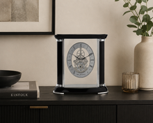 Load image into Gallery viewer, A black and silver mechanical clock with a transparent glass front, styled on a dark wooden console table with a vase, bowl, and framed art in a warm beige room.
