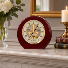 Load image into Gallery viewer, A round burgundy clock with a gold mechanical skeleton face sits on a marble mantelpiece in a styled home setting, flanked by a vase of white flowers and a lit candle on stacked books.

