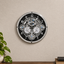 Load image into Gallery viewer, A round wall clock with a black face, silver-toned outer frame, and an intricate exposed mechanical gear and cogs design in the center. The clock is mounted on a textured beige wall above a wooden surface, with a green potted plant on the left and a stack of books on the right.
