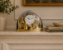 Load image into Gallery viewer, A decorative gold and black arched table clock with a miniature castle and architectural scene inside, displayed on a light marble mantel in a styled interior setting.
