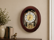 Load image into Gallery viewer, An oval wall clock with a dark reddish-brown wooden frame, featuring a cream-colored face and a detailed gold-toned clockwork mechanism visible behind glass. The clock is mounted on a beige wall above a shelf with books, a plant, and a small bird figurine.
