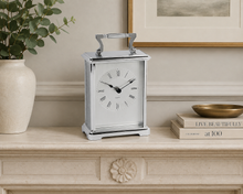 Load image into Gallery viewer, A polished silver metal mantel clock with a white face and black Roman numerals, styled on a beige stone console table next to a potted eucalyptus plant and decorative books.
