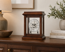 Load image into Gallery viewer, A wooden mantel clock with a glass front and visible inner gear mechanism, displayed on a dark wood sideboard in a styled room setting with a lamp, books, and a plant.
