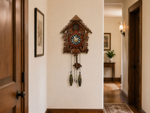 Load image into Gallery viewer, A traditional carved wooden cuckoo clock hangs on a cream-colored hallway wall, flanked by dark wood doors and a framed picture.
