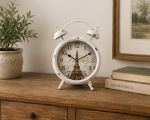 Load image into Gallery viewer, A white vintage-style twin-bell alarm clock with a Paris Tower Eiffel Tower design on the clock face, styled on a wooden cabinet next to a potted plant and books.
